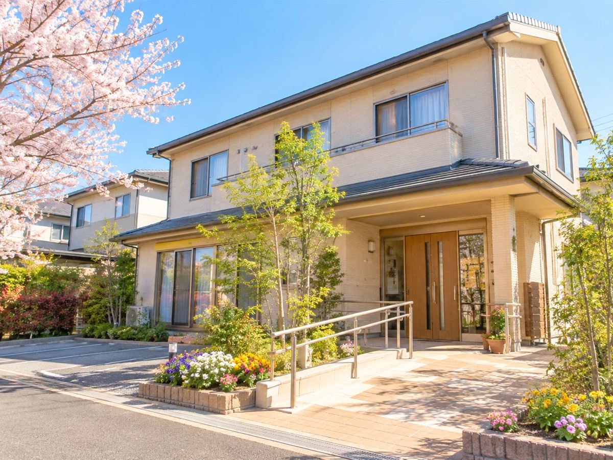 桜の木と花壇に囲まれた、2階建ての明るい建物の外観。玄関には手すり付きのスロープがある